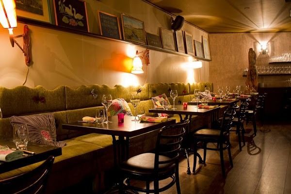A dimly lit restaurant interior with a green tufted booth, wooden tables, chairs, and framed artwork on the wall.
