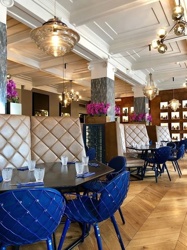 Upscale restaurant interior with tan tufted booths, blue circular chairs, dark wood tables, and decorative ceiling lights.