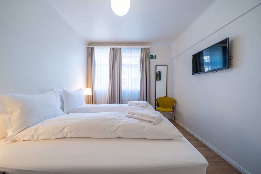 A hotel room featuring two twin beds joined together, white linens, a TV on the wall, and a small yellow chair by a mirror.