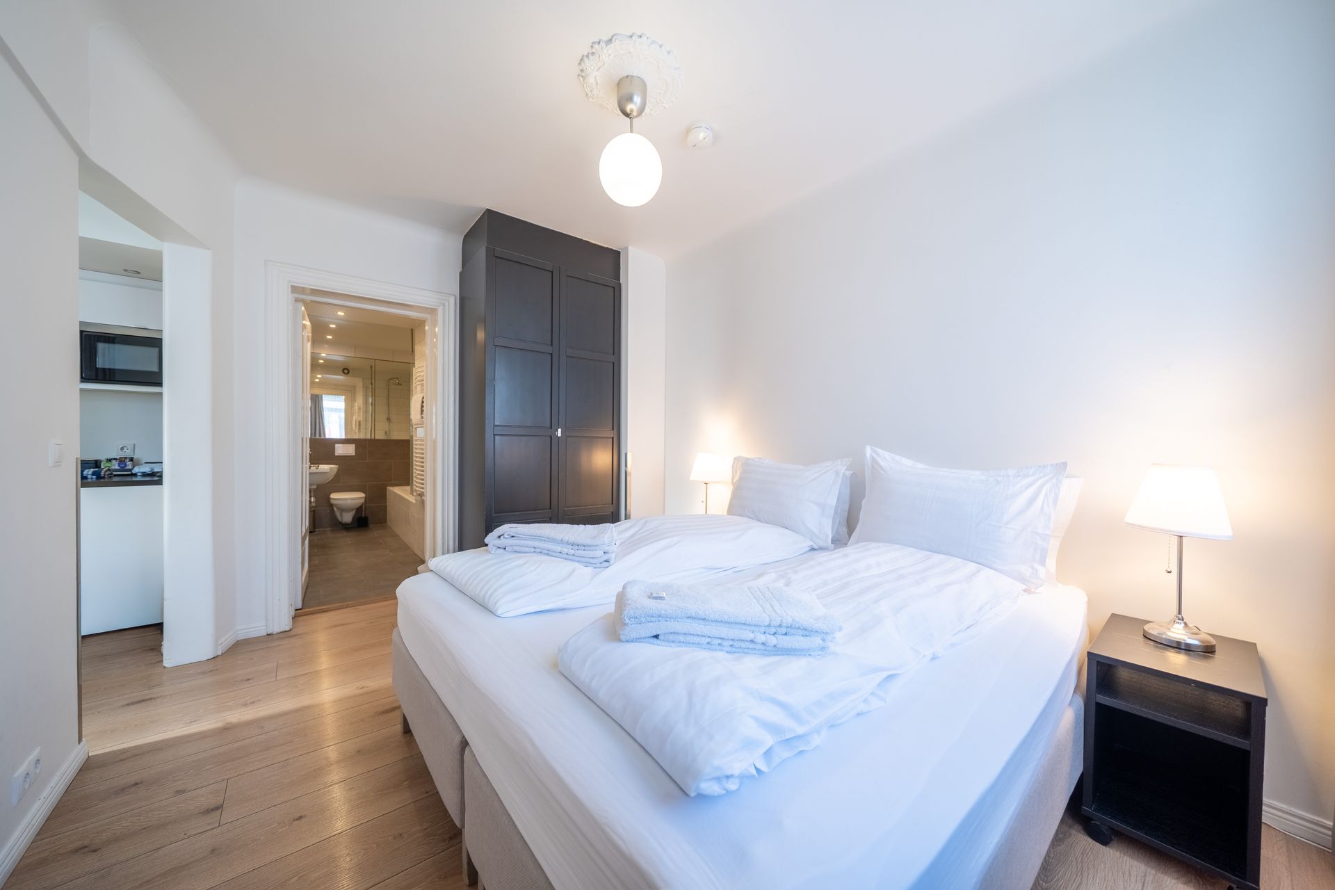 A hotel room with a neatly made double bed, dark wardrobe, bedside lamp, and an adjacent bathroom on a wood floor.
