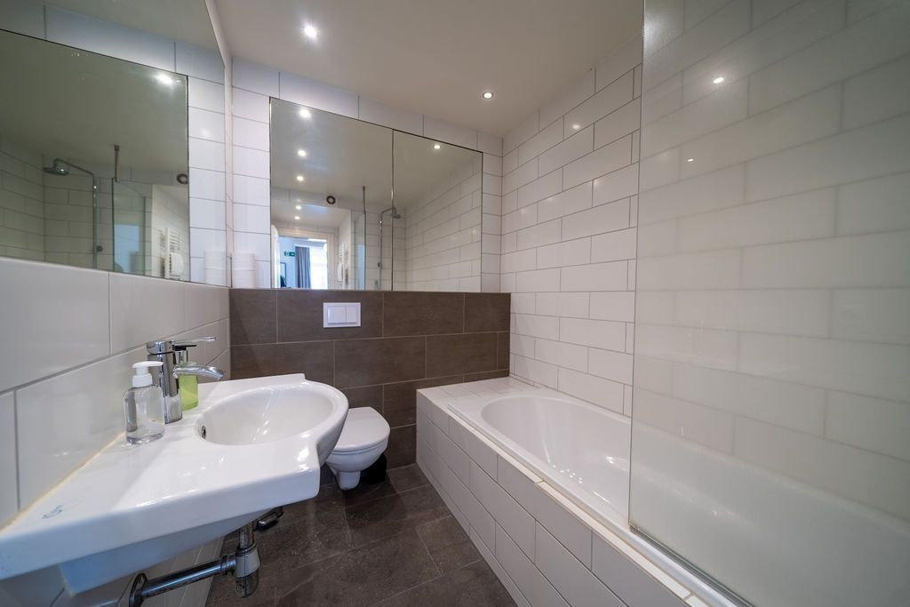A modern bathroom featuring a white pedestal sink, a toilet, a bathtub with a glass screen, and white subway-tiled walls.
