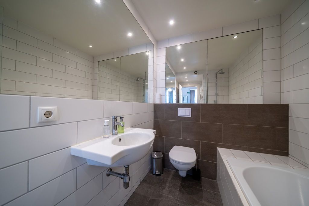 A modern bathroom with white subway tile walls, a white pedestal sink, a wall-mounted toilet, a bathtub, and mirrors.