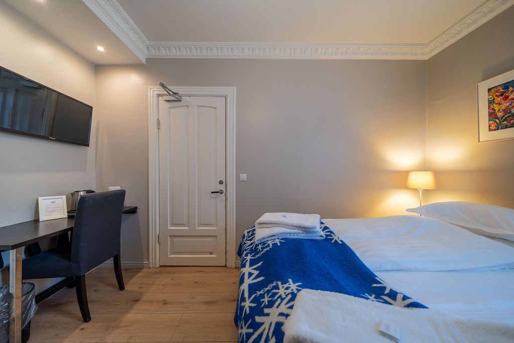 A hotel room featuring a bed with a blue patterned throw, a desk with a blue chair, and a closed white door.