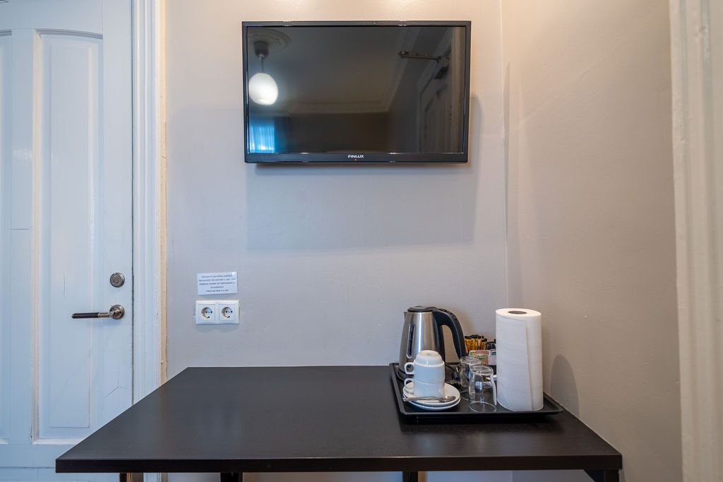 A wall-mounted TV above a small dark table with a kettle, tray, and paper towels in a room with a white door.