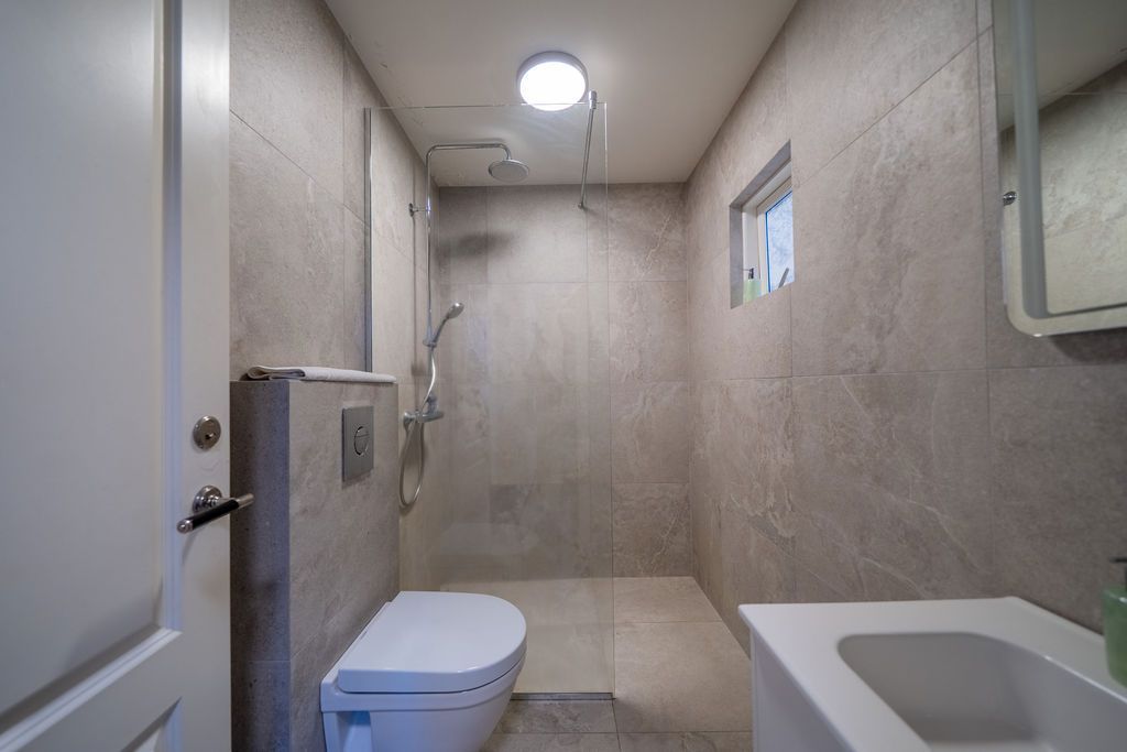 A modern bathroom featuring beige tiled walls, a wall-mounted toilet, a walk-in shower area, and a white sink.