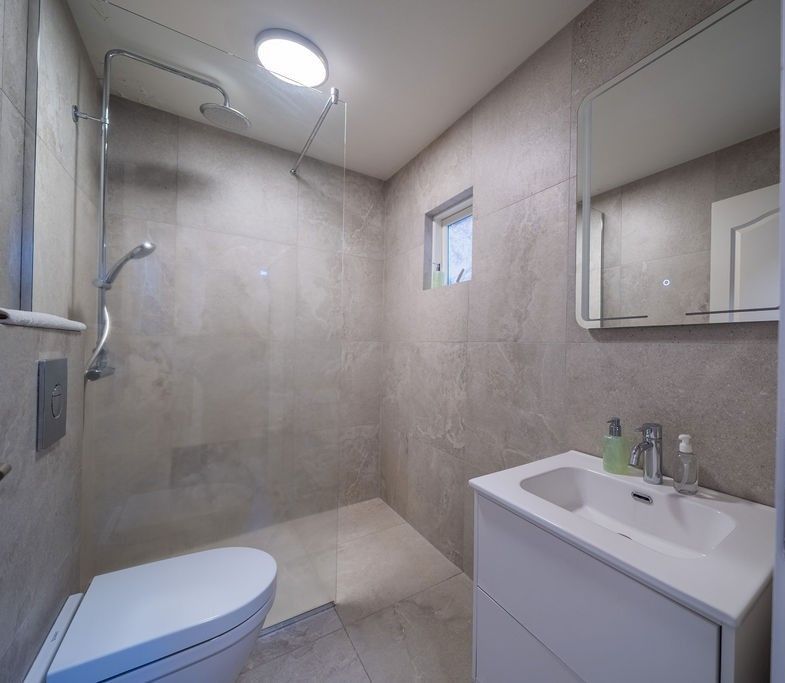 A modern, neutral-toned bathroom featuring a walk-in glass shower, wall-mounted toilet, and white vanity with a sink.
