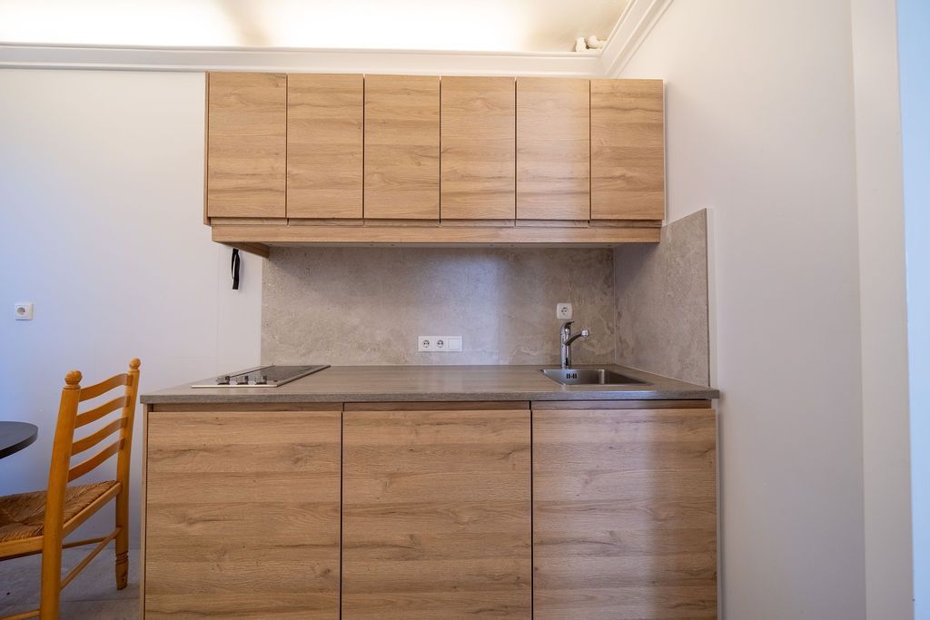 A small kitchen area with light wood-grain cabinets, a grey countertop, a stainless steel sink, and a cooktop.