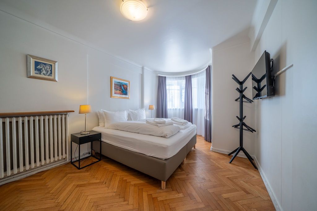 A bright, minimalist hotel room featuring a bed with white linens, a radiator, parquet floors, and a coat stand.
