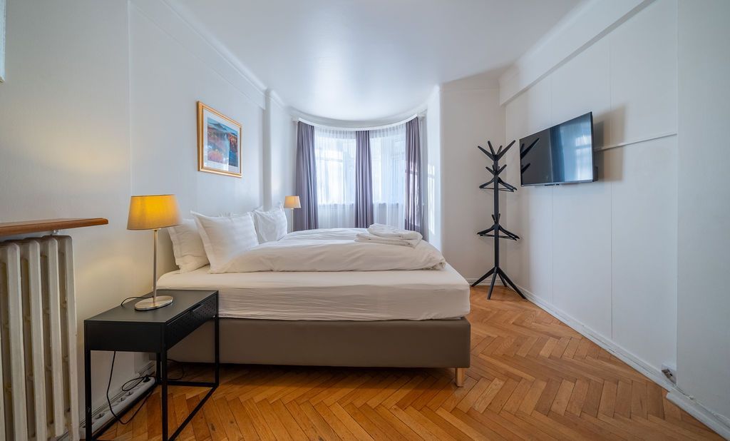 A bright, modern hotel room featuring a bed with white linens, a bedside table with a lamp, and a wall-mounted TV.