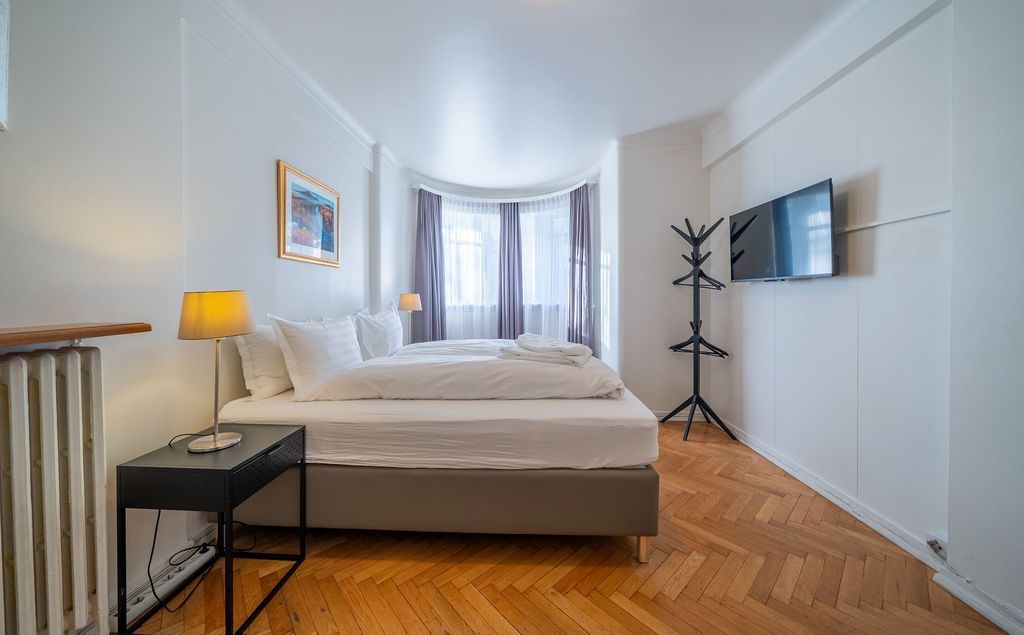 A minimalist hotel room with a bed, a bedside lamp on a side table, a wall-mounted TV, and a coat rack on wooden floors.