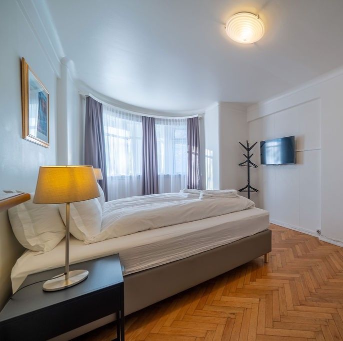 A bedroom with a bed, a nightstand with a lamp, a window with grey curtains, and a wall-mounted TV on a hardwood floor.