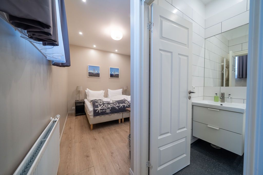 A bedroom with two beds and beige walls is visible through a doorway next to a bathroom with a white vanity and mirror.