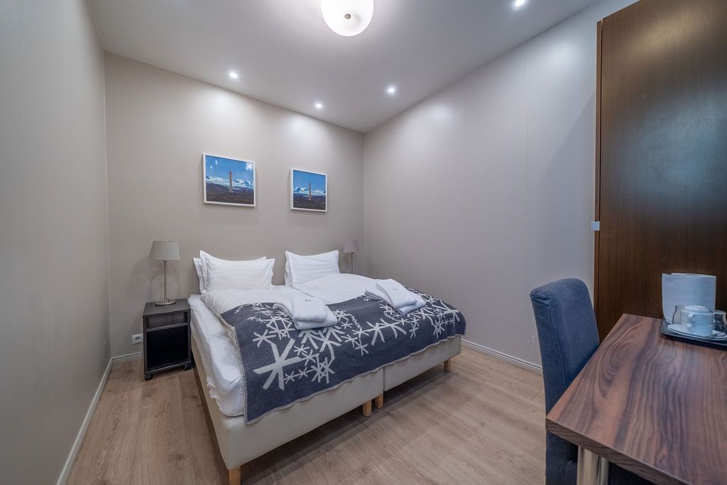 A modern hotel room with a bed featuring a snowflake-patterned throw, a desk with a chair, and two landscape paintings.