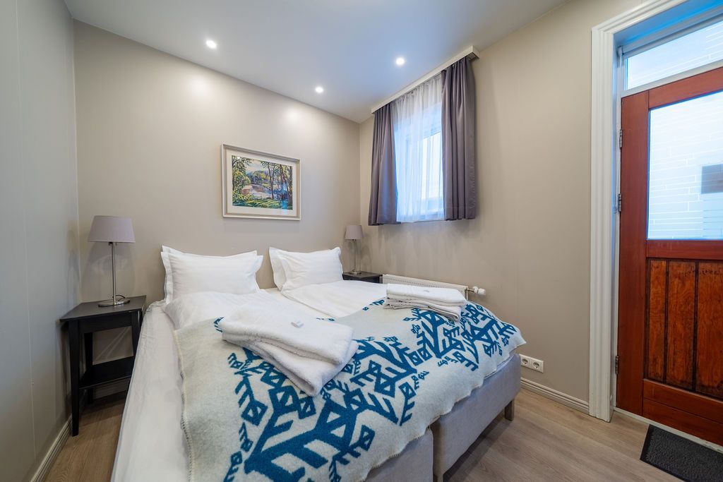 A clean, modern bedroom with a twin bed, white bedding, a blue patterned throw, a side table, and a wooden door.