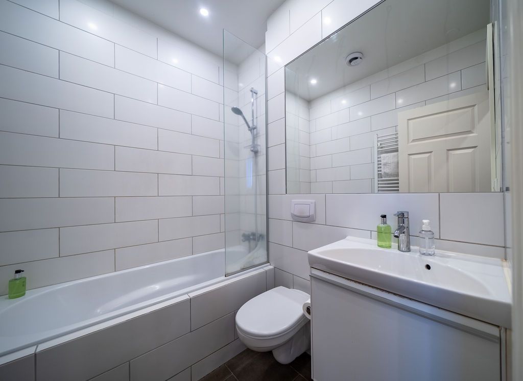A modern, bright bathroom with white tiled walls, a bathtub with a glass screen, a white toilet, and a vanity sink.