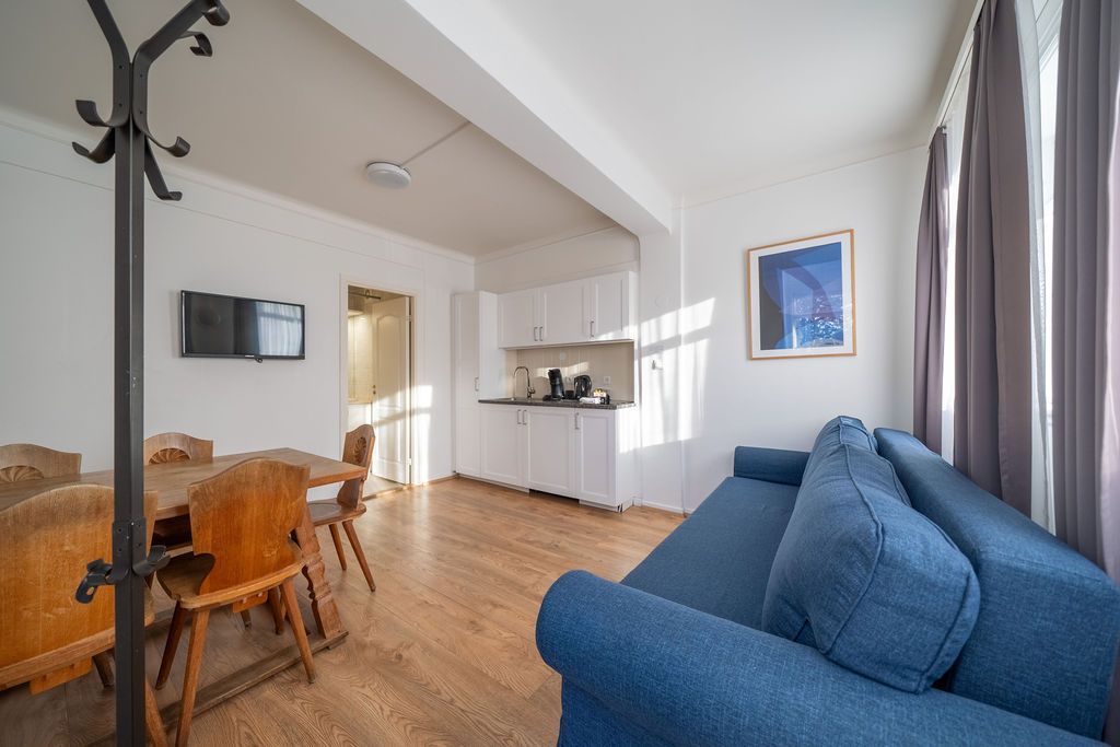 A furnished studio apartment with a dining table, kitchenette, and blue sofa in a brightly lit room with wood flooring.