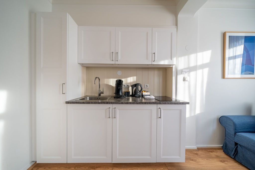 White kitchenette with upper and lower cabinets, a dark countertop, a sink, and kitchen appliances, next to a blue sofa.