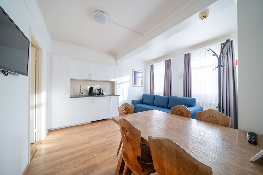 A bright hotel studio with light wood flooring, a kitchenette, a large wooden dining table, and a blue sofa.