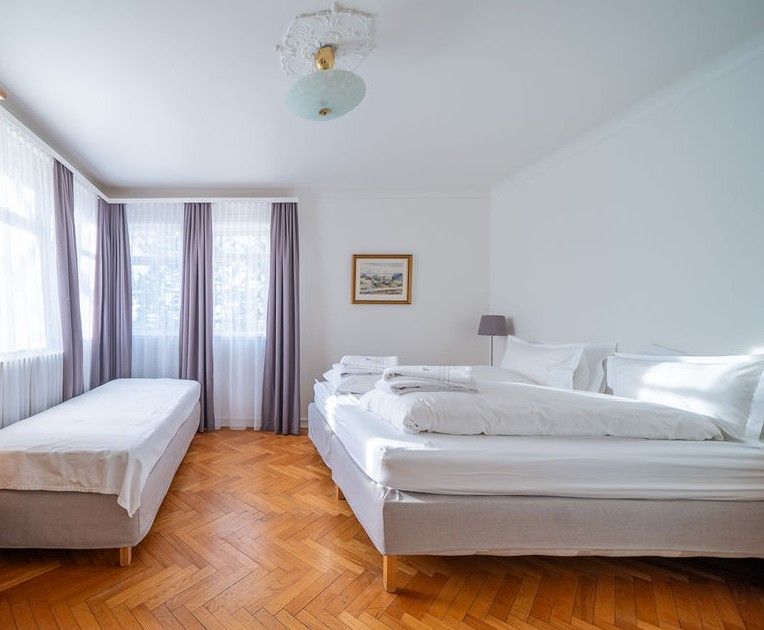 A bedroom with herringbone wood floors, a twin bed on the left, a double bed on the right, and grey curtains.