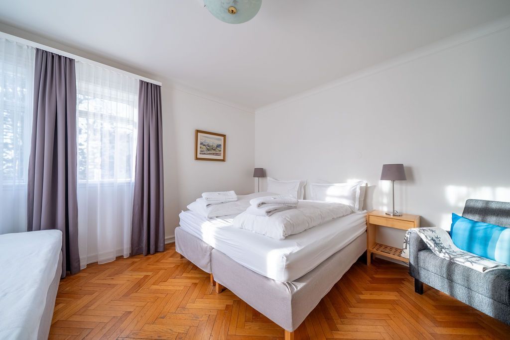 A bedroom with two twin beds featuring white bedding, a gray armchair, wooden parquet floors, and purple-gray curtains.