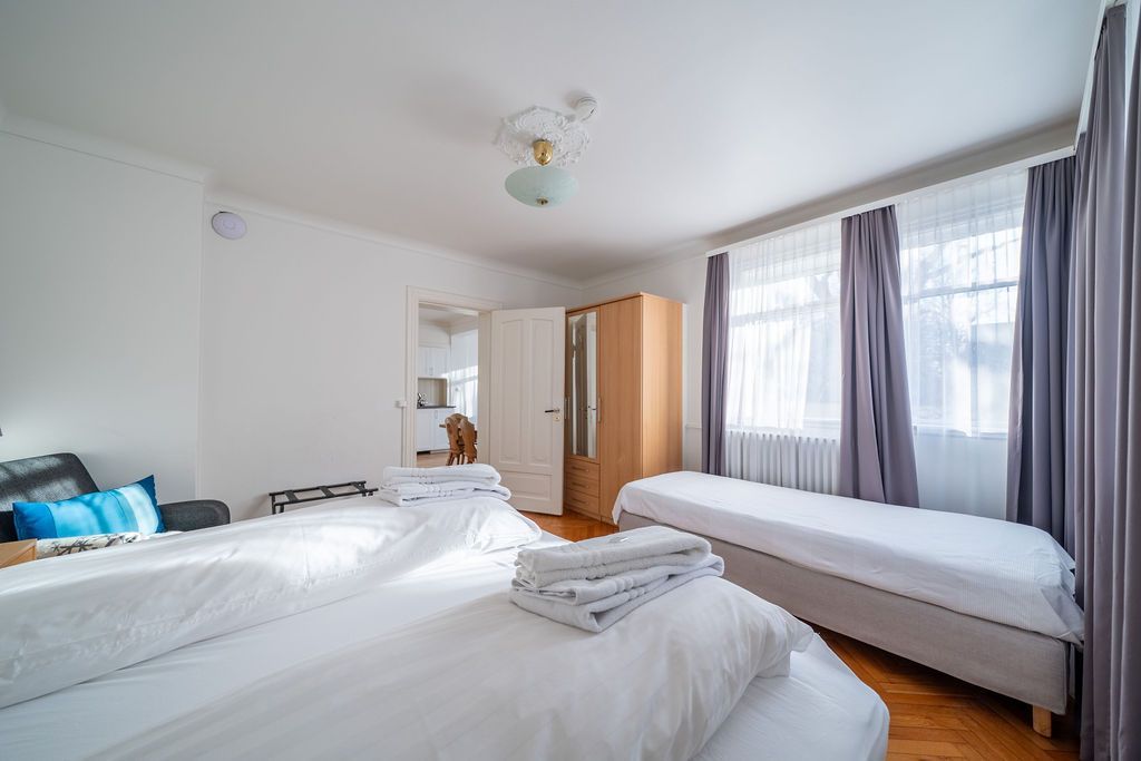 A bedroom with a large double bed, a separate single bed by the window with grey curtains, and a wooden wardrobe.