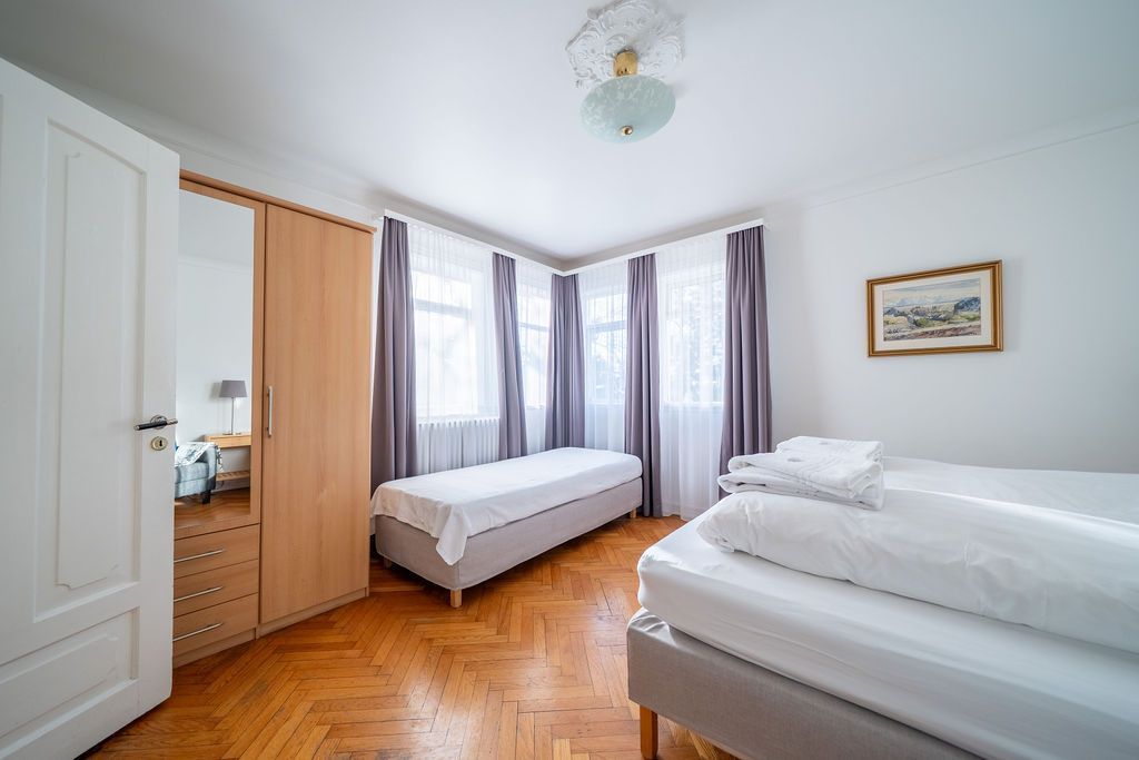 A bedroom with two beds, a wooden wardrobe with a mirror, light-colored walls, wood floors, and gray floor-length curtains.