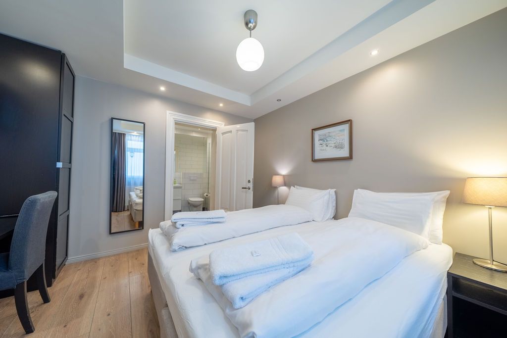A tidy hotel room featuring two twin beds with white linens, a mirror, and a small desk lamp on a light wood floor.