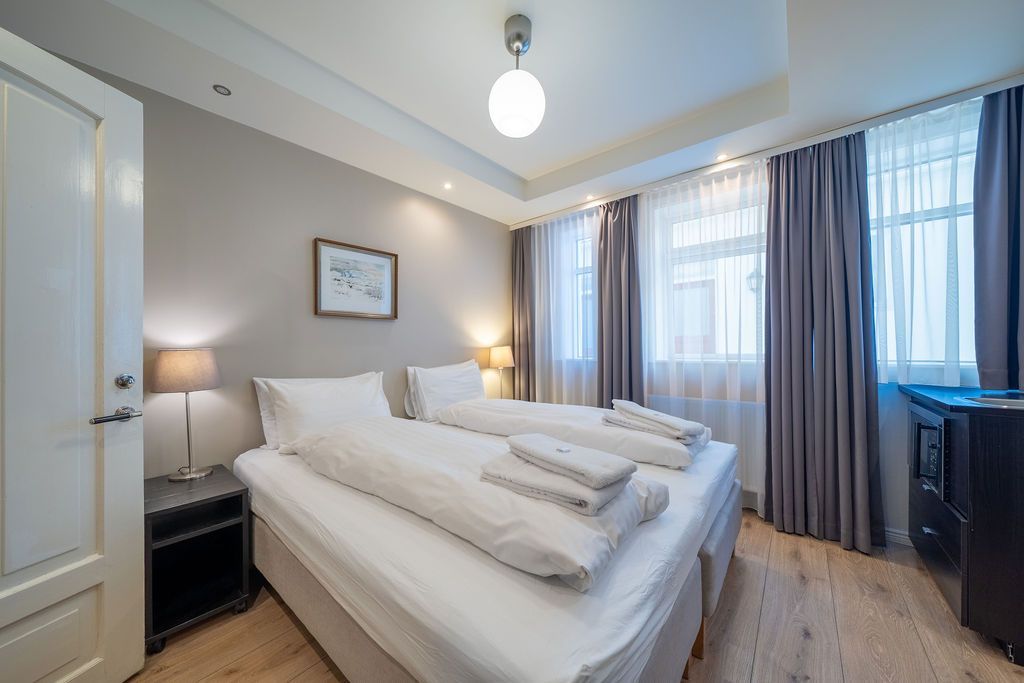 A hotel room with two twin beds featuring white bedding, light gray walls, a nightstand, and floor-to-ceiling gray curtains.
