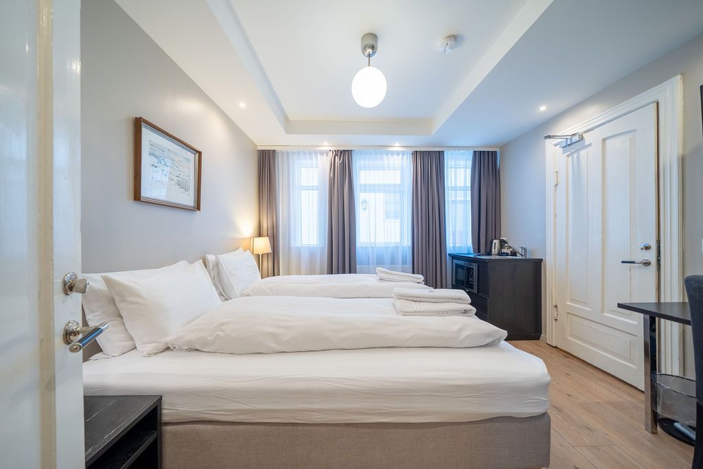 A hotel room with two white beds, light gray walls, dark curtains over a window, and a white door on the right.