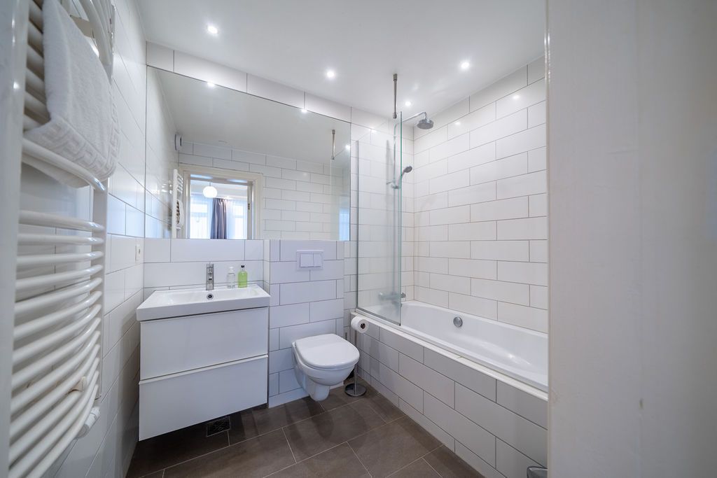 A white bathroom with a vanity, toilet, and bathtub tiled in a white, patterned design, illuminated by ceiling spotlights.