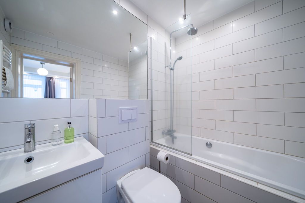 A clean, modern bathroom with a white vanity sink, a toilet, and a bathtub with a glass shower screen and white tile walls.
