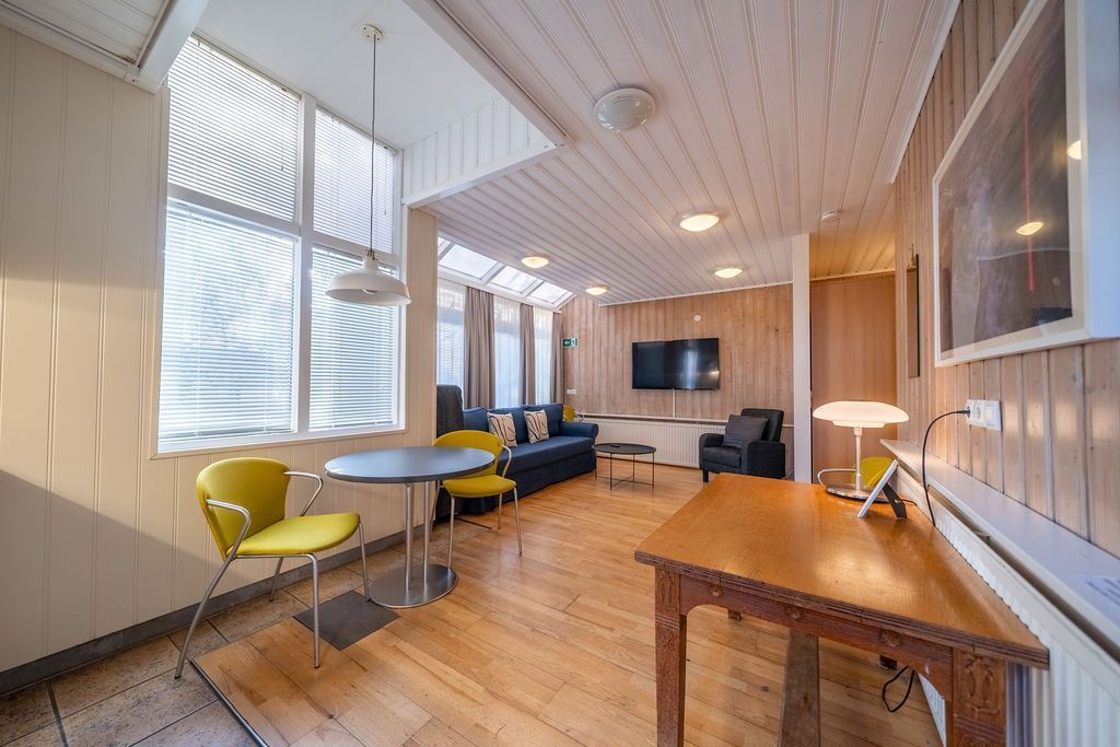 A bright living area with wooden paneling, a circular dining table with yellow chairs, a blue sofa, and a wood desk.