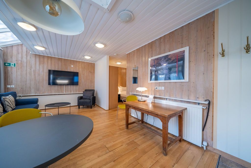 A bright lounge with wooden walls, a round table, a blue sofa, a wooden desk, and a wall-mounted TV.