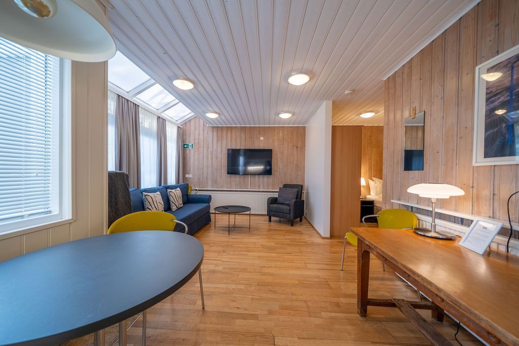 A modern hotel suite with wood paneling, featuring a dining table, a blue sofa, an armchair, a TV, and a desk area.