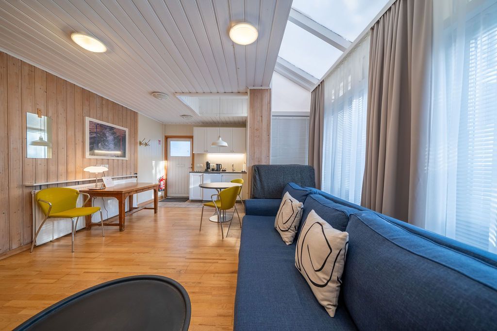 A brightly lit modern living room with a blue sofa, wood paneled walls, a dining area, and large windows with curtains.