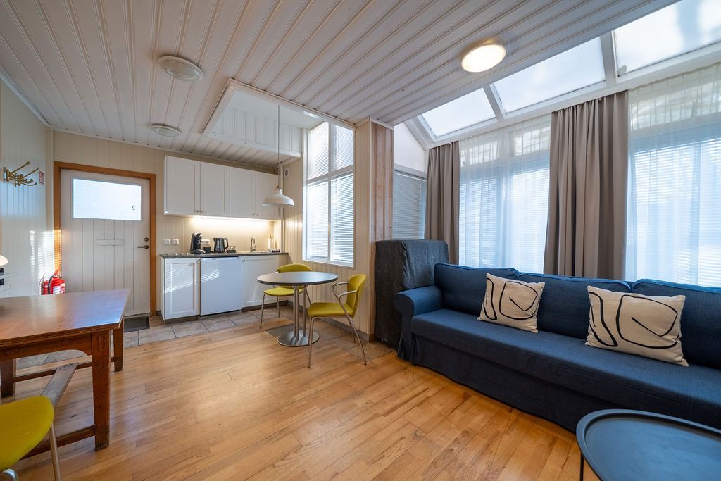 A sunroom with a blue sofa, a dining table, a small kitchen area, and a large window wall with floor-length curtains.