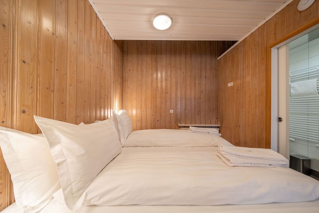 A small, modern hotel cabin with wooden paneling walls, two neatly made white beds, and a glass-paneled bathroom entrance.