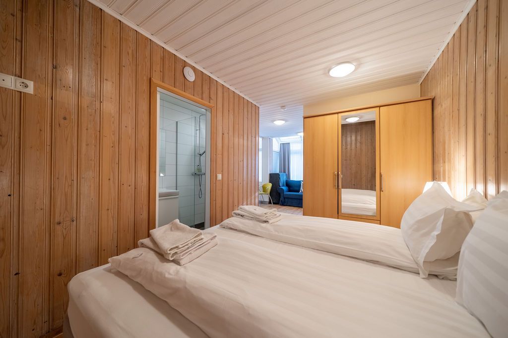 A bedroom featuring two twin beds, light wood-paneled walls, a wooden wardrobe with a mirror, and a bathroom doorway.