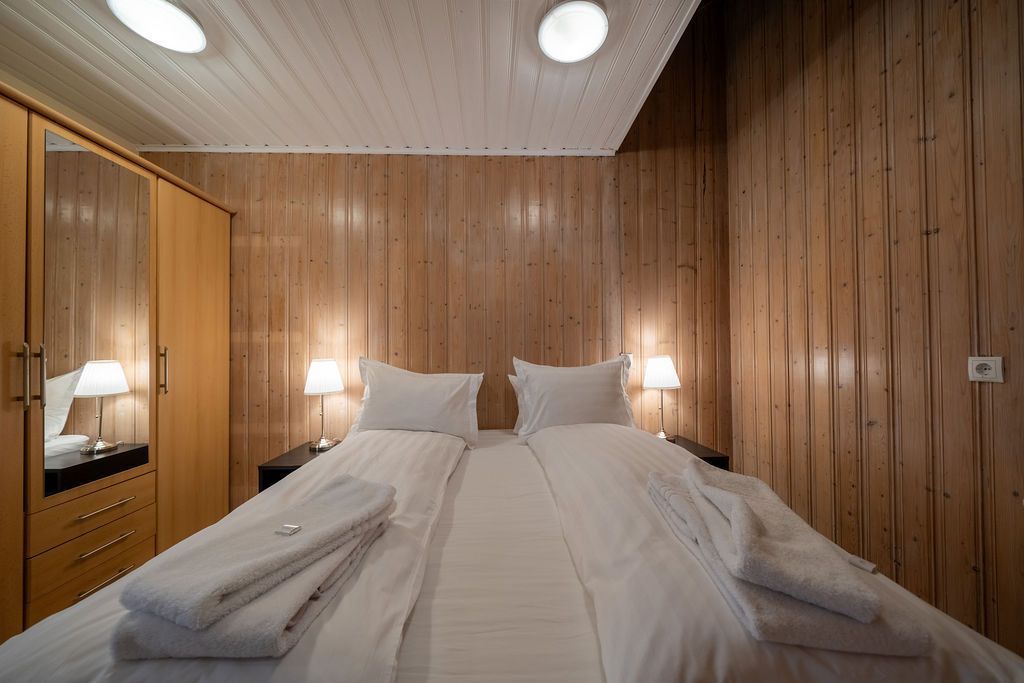 A double bed with white bedding and folded towels in a room with light wood paneled walls and ceiling, and a wooden wardrobe.