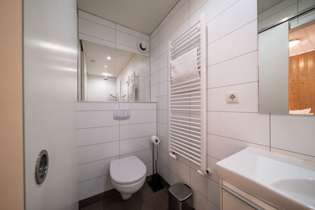 Modern, white-tiled bathroom featuring a toilet, a wall-mounted towel heater, a mirror, and a small sink.