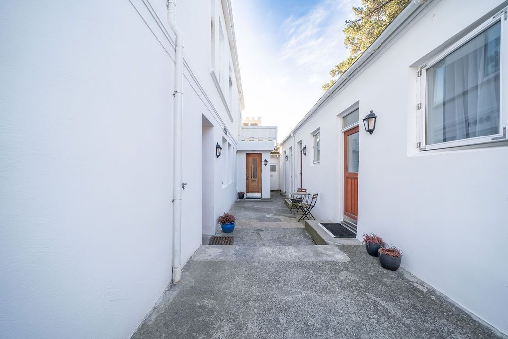 A narrow alley between two white buildings with brown wooden doors, leading to a distant structure under a blue sky.