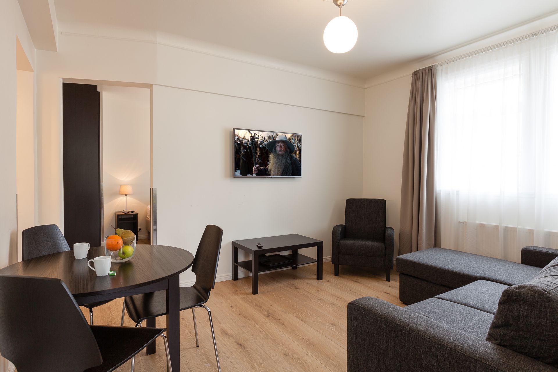 A modern apartment living room with a gray sectional, dark dining set, television, and light wood flooring.