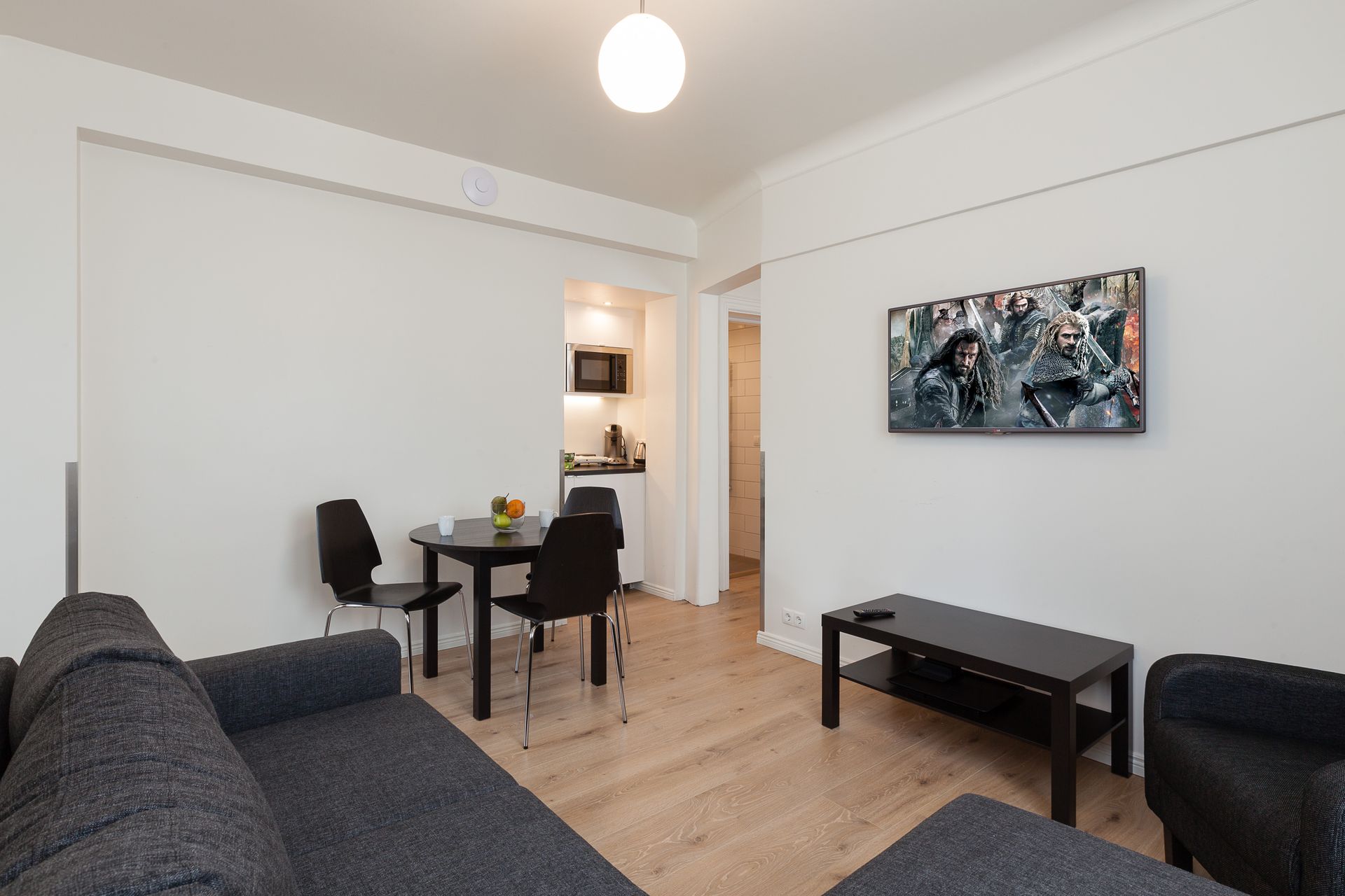A minimalist living room with a gray sofa, a small dining table with black chairs, and a wall-mounted TV.