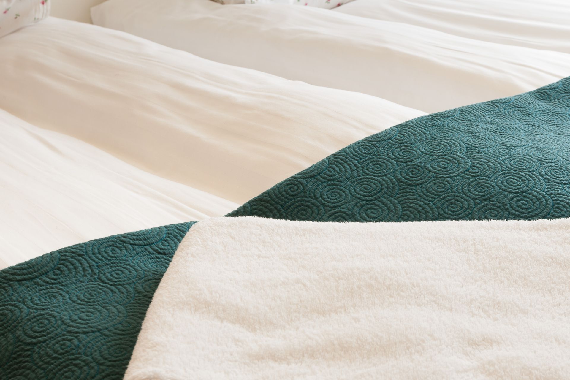 A white bed with a textured, deep teal blanket folded over the foot, partially covered by a light beige throw blanket.
