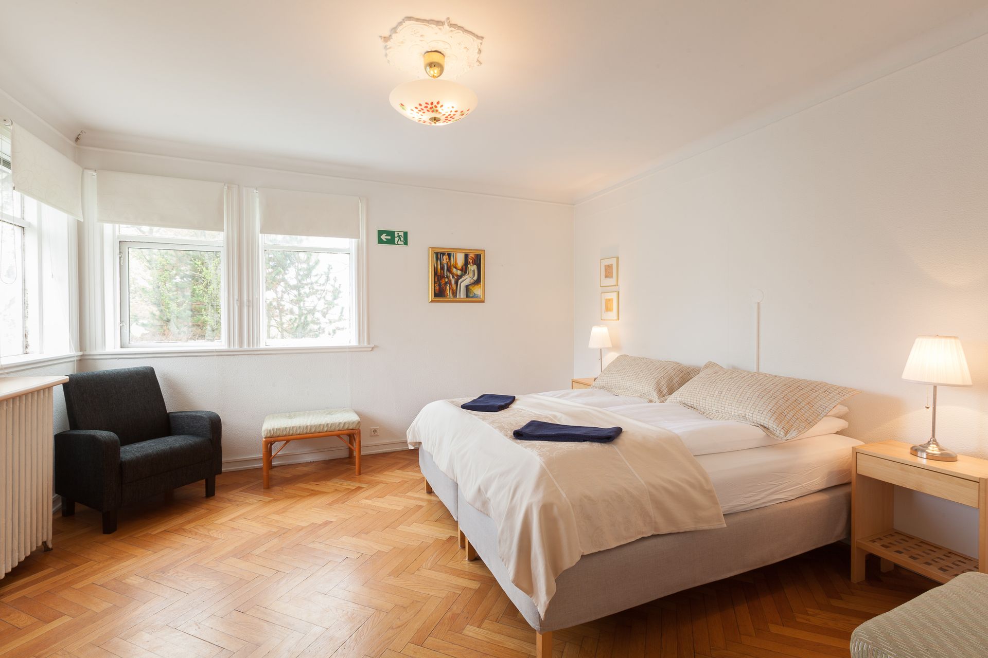 A bright hotel bedroom with a large double bed, an armchair, a small side table, and parquet flooring.