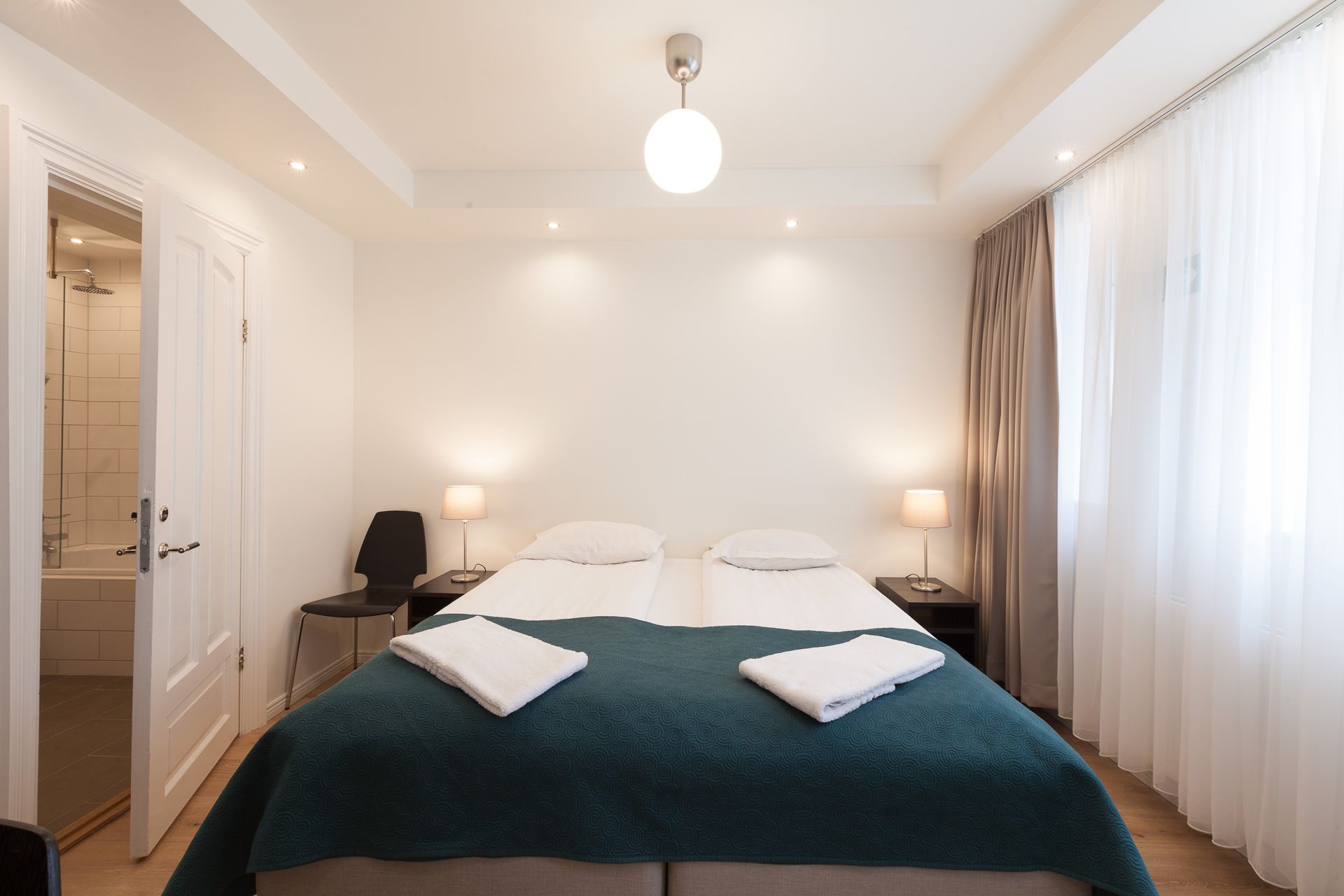 A bright bedroom with a queen bed featuring a dark teal quilt, two nightstands with lamps, a side chair, and an open door.