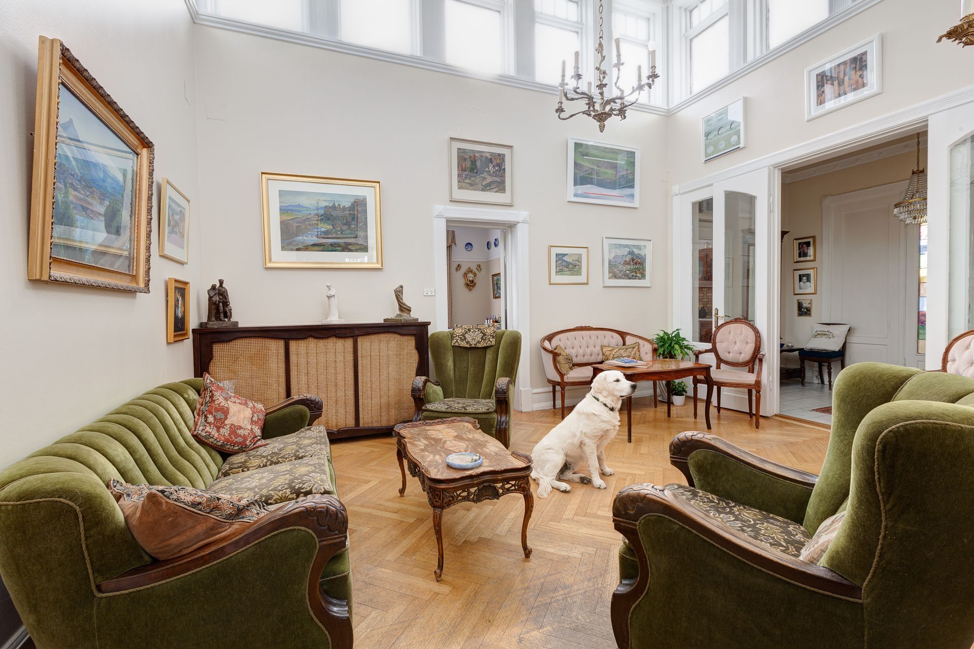 A bright, formal living room with green velvet sofas, wooden furniture, artwork on the walls, and a sitting golden dog.