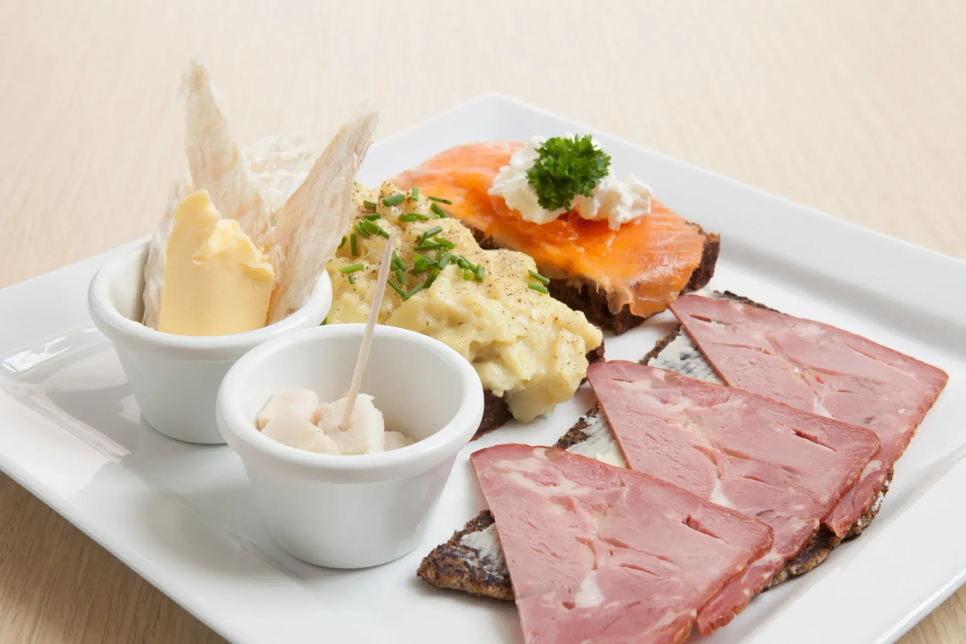 A platter of open-faced smørrebrød sandwiches with sliced meat, fish, and potato salad, served with butter and spreads.