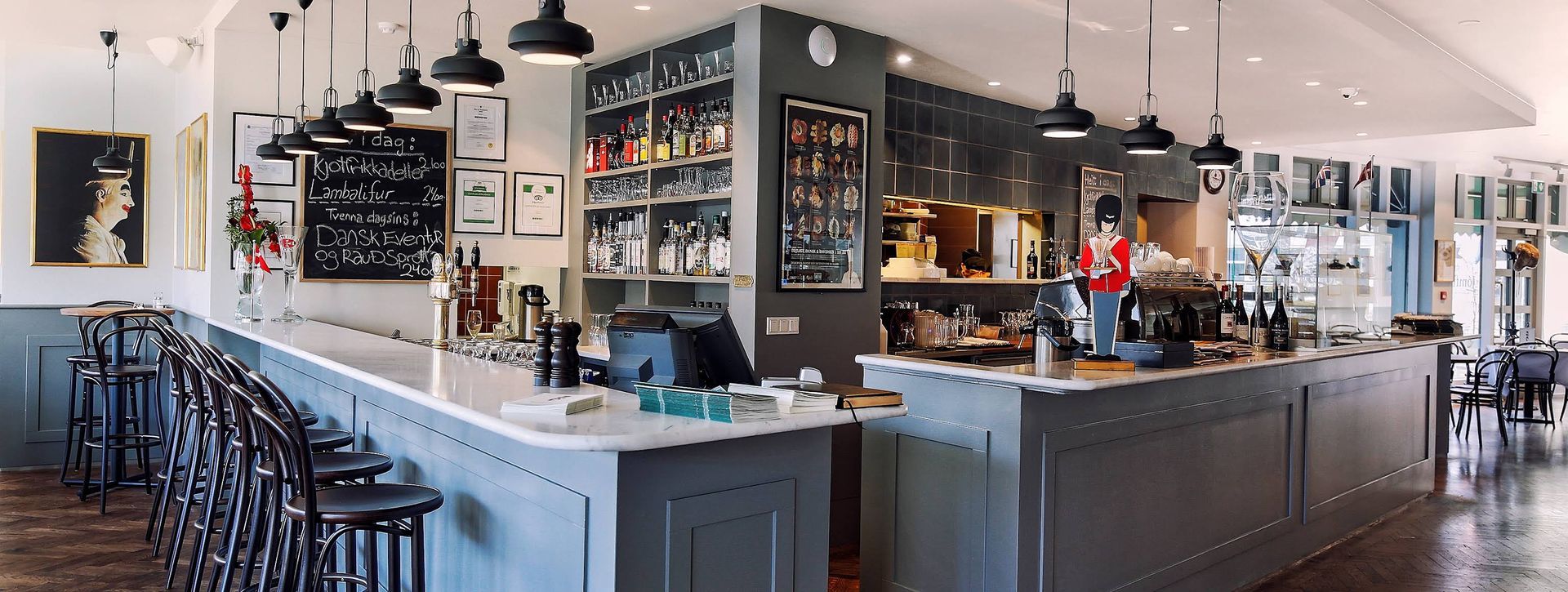 A modern bar interior with light-colored countertops, dark gray cabinetry, hanging pendant lights, and stools.