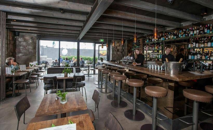 A modern restaurant interior with a long bar, stools, wooden tables, and large windows looking out onto an outdoor patio.